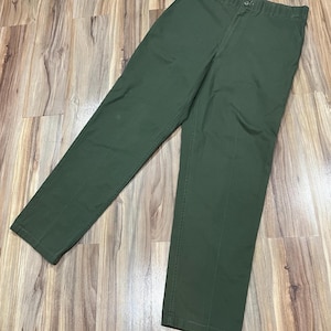 May include: Olive green pants with a button closure and pockets. The pants are made of a durable fabric and have a classic, straight-leg design. The pants are laid flat on a wooden surface.
