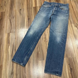 May include: A pair of blue denim jeans featuring a classic five-pocket design, a button fly closure, and a faded wash with whiskering details on the thighs. The jeans have a straight-leg cut with visible stitching along the seams and pockets.