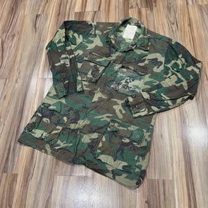 May include: A long-sleeved camouflage jacket featuring a woodland pattern in shades of green, brown, and black. The jacket includes a pointed collar, a button-front closure, four large front pockets with buttoned flaps, and a white label at the neck.
