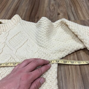 May include: A cream-colored knitted sweater with a cable knit pattern. A measuring tape is placed across the sweater, showing a measurement of 25 inches.
