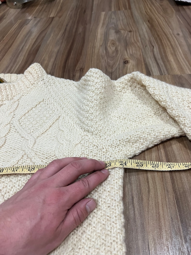 May include: A cream-colored knitted sweater with a cable knit pattern. A measuring tape is placed across the sweater, showing a measurement of 25 inches.