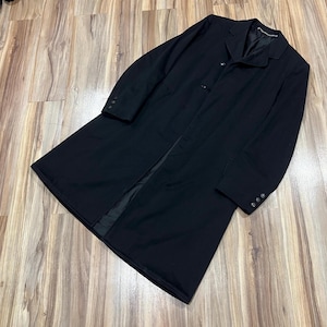 May include: A long, black coat with a classic design. The coat features a notched lapel, three buttons, and buttoned cuffs. The coat is made of a smooth, dark fabric and is laid out on a wooden surface.