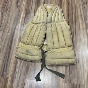 May include: A vintage, quilted vest in a faded olive green color. The vest has a padded design with vertical stitching and a front closure. It features a label at the neckline and olive green ties. The vest appears worn, suggesting its age and use.