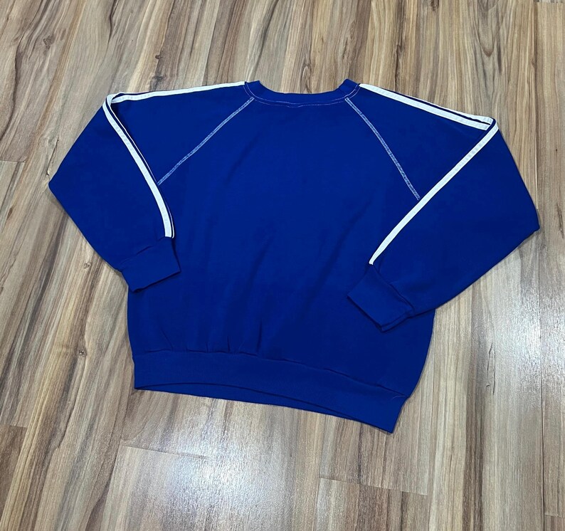May include: A royal blue sweatshirt with white stripes on the sleeves. The sweatshirt has a crew neck and a relaxed fit.