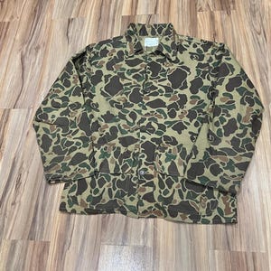 Puede incluir: Chaqueta de camuflaje en tonos marrones, verdes y beige. La chaqueta tiene cierre de botones, dos bolsillos delanteros y cuello. El patrón es un diseño clásico de camuflaje, adecuado para actividades al aire libre. La chaqueta es de talla mediana.
