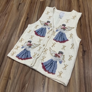 May include: Cream-colored, button-down vest with embroidered angel designs. The angels have gold wings, red and brown hair, and blue dresses with red trim. Gold star and cross accents are also embroidered.