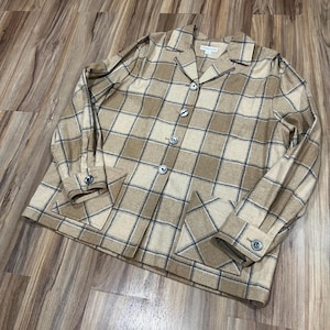 May include: A tan and beige plaid button-down shirt with a collar, long sleeves with buttoned cuffs, and two large square front pockets. The shirt features a Pendleton brand label at the inner neckline and dark buttons down the center front.