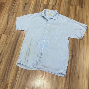 May include: A light blue short-sleeved button-down shirt with a chest pocket. The shirt has a classic collar and white buttons. The fabric appears to be slightly wrinkled, suggesting a casual style. The shirt is laid flat on a wooden surface.
