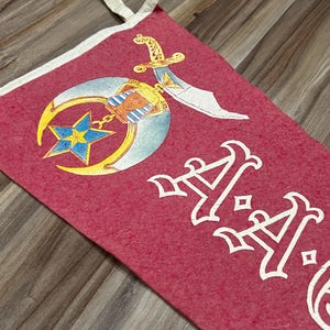 May include: Red pennant with white trim and a white emblem. The emblem includes a crescent moon, star, and sword. The pennant also features white text. The pennant is made of a textured material.