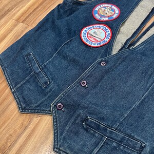 May include: A blue denim vest with two embroidered patches. The patches are circular and feature the text "1989 Daytona 500 World Center of Racing" and "30th Annual Daytona 500 1959-1988".