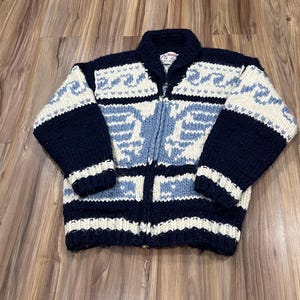 May include: A navy blue, cream, and light blue zip-up sweater with a geometric pattern. The sweater features a high collar, long sleeves, and a full zipper. The design includes a repeating pattern of geometric shapes and lines.