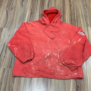 May include: A red hooded sweatshirt featuring a front kangaroo pocket, long sleeves, and a drawstring hood. The fabric has a distressed appearance with white paint splatters throughout and a small Marlboro logo patch on the left sleeve.