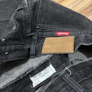 Puede incluir: Primer plano de unos vaqueros Wrangler negros. Los vaqueros tienen una etiqueta roja de Wrangler y un parche de cuero marr&oacute;n. Se ve una etiqueta blanca con el logotipo de Wrangler y la informaci&oacute;n de la talla. Los vaqueros son 100% algod&oacute;n.