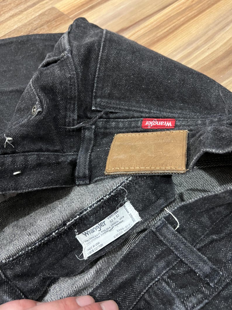 Puede incluir: Primer plano de unos vaqueros Wrangler negros. Los vaqueros tienen una etiqueta roja de Wrangler y un parche de cuero marr&oacute;n. Se ve una etiqueta blanca con el logotipo de Wrangler y la informaci&oacute;n de la talla. Los vaqueros son 100% algod&oacute;n.