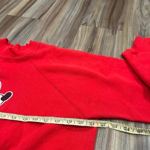 May include: A red sweatshirt with a white and black graphic on the front. A measuring tape is laid across the sleeve, showing a measurement of approximately 24 inches. The sweatshirt is made of a soft material.