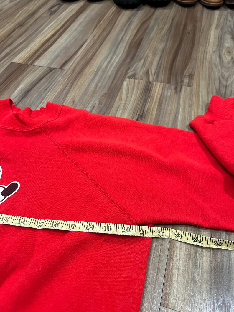 May include: A red sweatshirt with a white and black graphic on the front. A measuring tape is laid across the sleeve, showing a measurement of approximately 24 inches. The sweatshirt is made of a soft material.