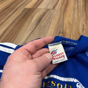 May include: A blue and white striped t-shirt with a yellow and white graphic of the state of Minnesota. The tag reads "Bassett Walker" and "Made in U.S.A."