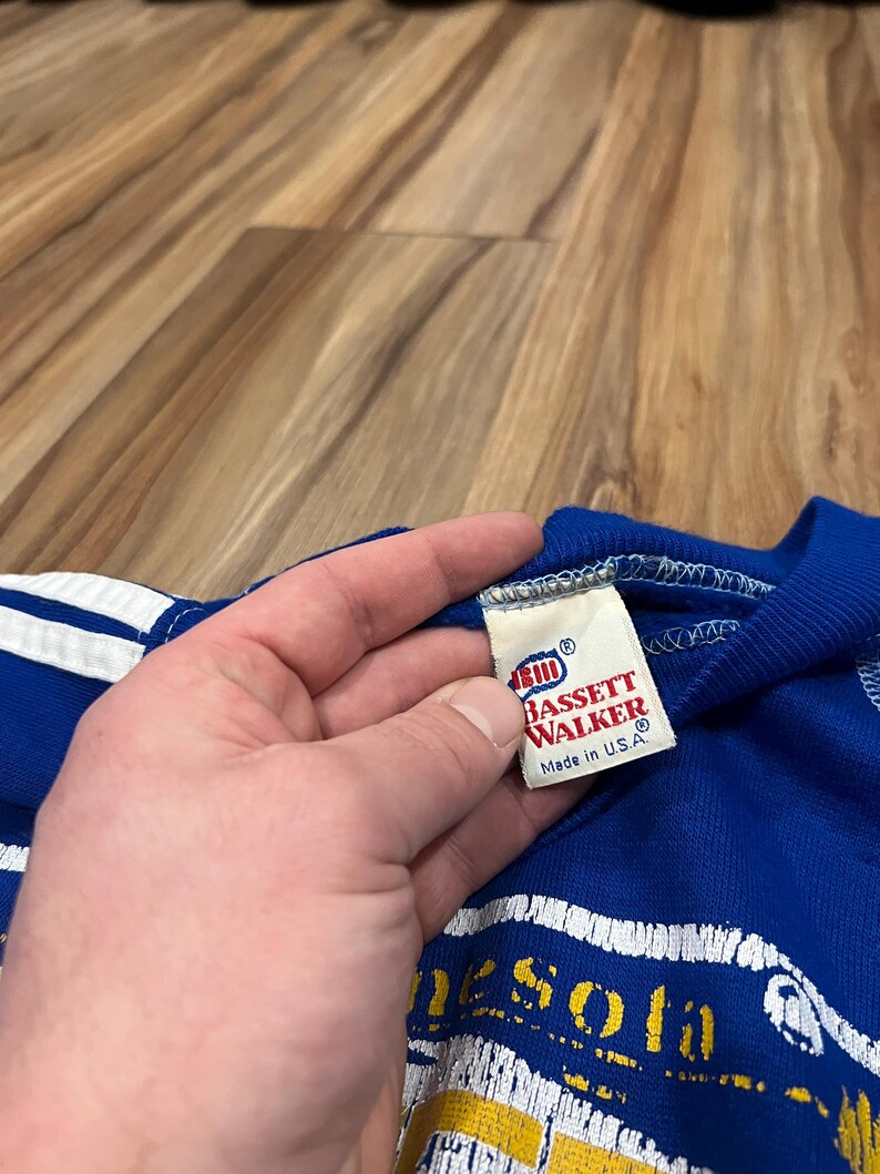 May include: A blue and white striped t-shirt with a yellow and white graphic of the state of Minnesota. The tag reads "Bassett Walker" and "Made in U.S.A."