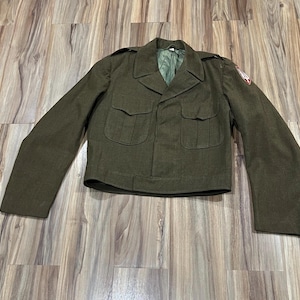 May include: An olive drab wool military-style jacket featuring a notched collar, long sleeves, and two front chest pockets with buttoned flaps. A red and white embroidered patch is visible on the left shoulder, and the interior is lined.