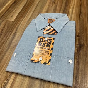 May include: A light blue long-sleeve shirt with a white button-down collar. The shirt has a pocket on the left chest and a tag that reads "BIG BEN by Wrangler Chambray Shirt".