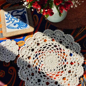 May include: A handmade, off-white crochet doily with a matching coaster sits on a patterned fabric. A small, square tile with a blue fish design and a vase of red flowers are also present. The doily has a detailed, openwork design.