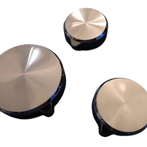 May include: Three silver-toned knobs with a circular design. The knobs have a brushed metal finish on the top and a black base with a small handle. The knobs are likely for a stove or other appliance.