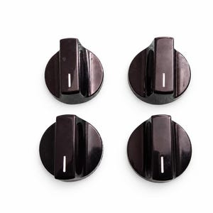 May include: A set of four circular black control knobs, each featuring a raised central grip and a single vertical white indicator line. These glossy plastic components are arranged in a two-by-two grid pattern against a plain white background.