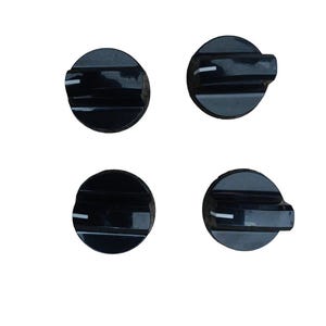 May include: A set of four circular black control knobs, each featuring a raised horizontal ridge and a single white indicator line on the top surface. These knobs are designed for appliance operation and have a smooth, glossy finish throughout.