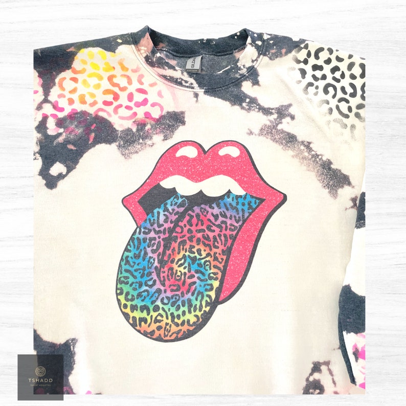 K&ouml;nnte beinhalten: Ein wei&szlig;es T-Shirt mit einem schwarzen und wei&szlig;en Batik-Muster. Das Shirt zeigt eine gro&szlig;e, farbenfrohe Grafik einer Zunge, die aus einem Mund herausragt. Die Zunge ist mit einem Leopardenmuster bedeckt.