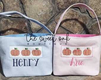 May include: Two personalised pumpkin trick-or-treat bags. One is blue with the name "Henry" and the other is pink with the name "Aria". Both bags feature a row of three embroidered pumpkins.