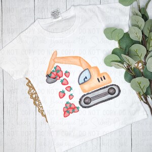 以下が含まれることがあります： 白いTシャツに、イチゴを掘っている黄色のショベルカーが描かれています。ショベルカーは白い背景にあり、その周りに「Sweet Oak Boutique the Sweet Oak」という文字が繰り返し書かれています。