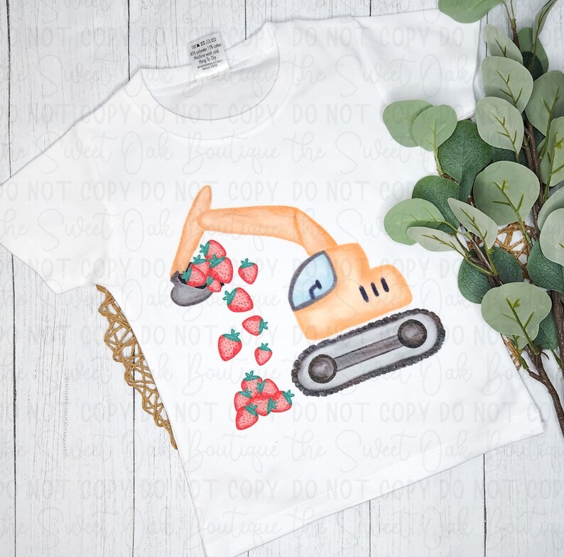以下が含まれることがあります： 白いTシャツに、イチゴを掘っている黄色のショベルカーが描かれています。ショベルカーは白い背景にあり、その周りに「Sweet Oak Boutique the Sweet Oak」という文字が繰り返し書かれています。