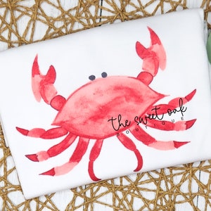 May include: A white t-shirt with a red watercolor crab illustration. The crab has two black eyes and is in a relaxed pose. The text "the sweet oak boutique" is printed in black below the crab.
