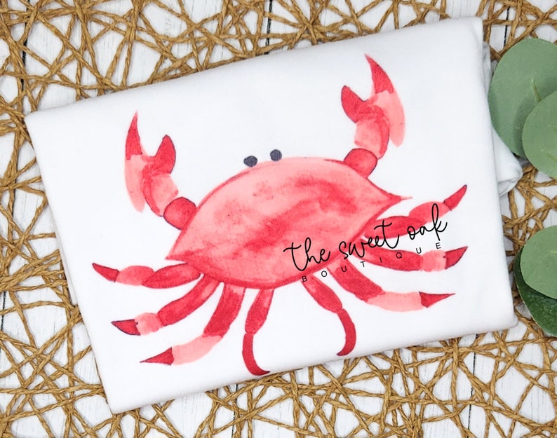 May include: A white t-shirt with a red watercolor crab illustration. The crab has two black eyes and is in a relaxed pose. The text "the sweet oak boutique" is printed in black below the crab.