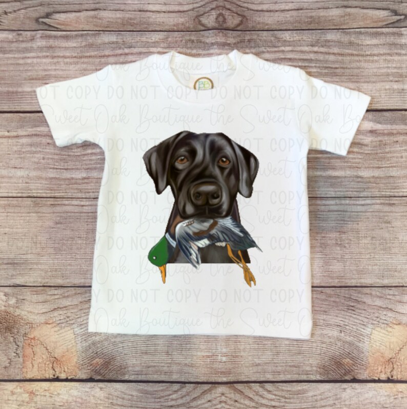 May include: White t-shirt with a black Labrador Retriever graphic carrying a duck. The dog and duck are realistically depicted. The shirt has the text "Sweet Oak Boutique" as a watermark.
