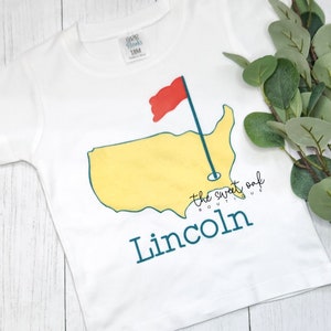 May include: White baby t-shirt with a yellow outline of the United States, a red golf flag, and a green golf club. The text "the sweet oak boutique" and "Lincoln" are printed in blue.