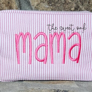 May include: Pink and white striped cosmetic bag with the word "mama" embroidered in pink. The bag has a handle on the side and the text "the sweet oak boutique" above the word "mama".