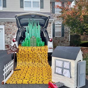 May include: A Halloween display featuring a car trunk transformed into the Emerald City from The Wizard of Oz. Includes a yellow brick road, a small house, and a sign that reads "Haunted Forest, Witches Castle, Mile, Turn Back If Were You!"