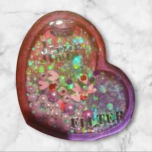 May include: A heart-shaped resin keychain with a glittery, iridescent background. The keychain features pink and white beads, small pink hearts, and the words "Jimin Filter" in silver lettering.