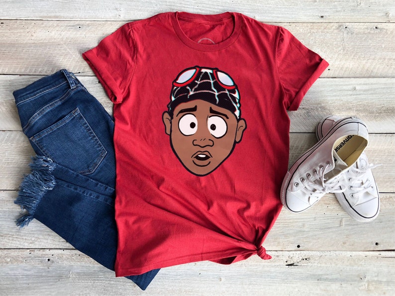 Pode incluir: Camiseta vermelha com uma ilustra&ccedil;&atilde;o de desenho animado de uma pessoa usando um chap&eacute;u vermelho e preto e &oacute;culos de prote&ccedil;&atilde;o. A pessoa tem pele marrom e parece surpresa.