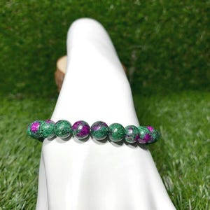 May include: A beaded bracelet featuring round, polished stones with a mottled appearance of deep magenta and forest green. The beads are strung together in a single row, showcasing a natural color variation throughout each individual stone.