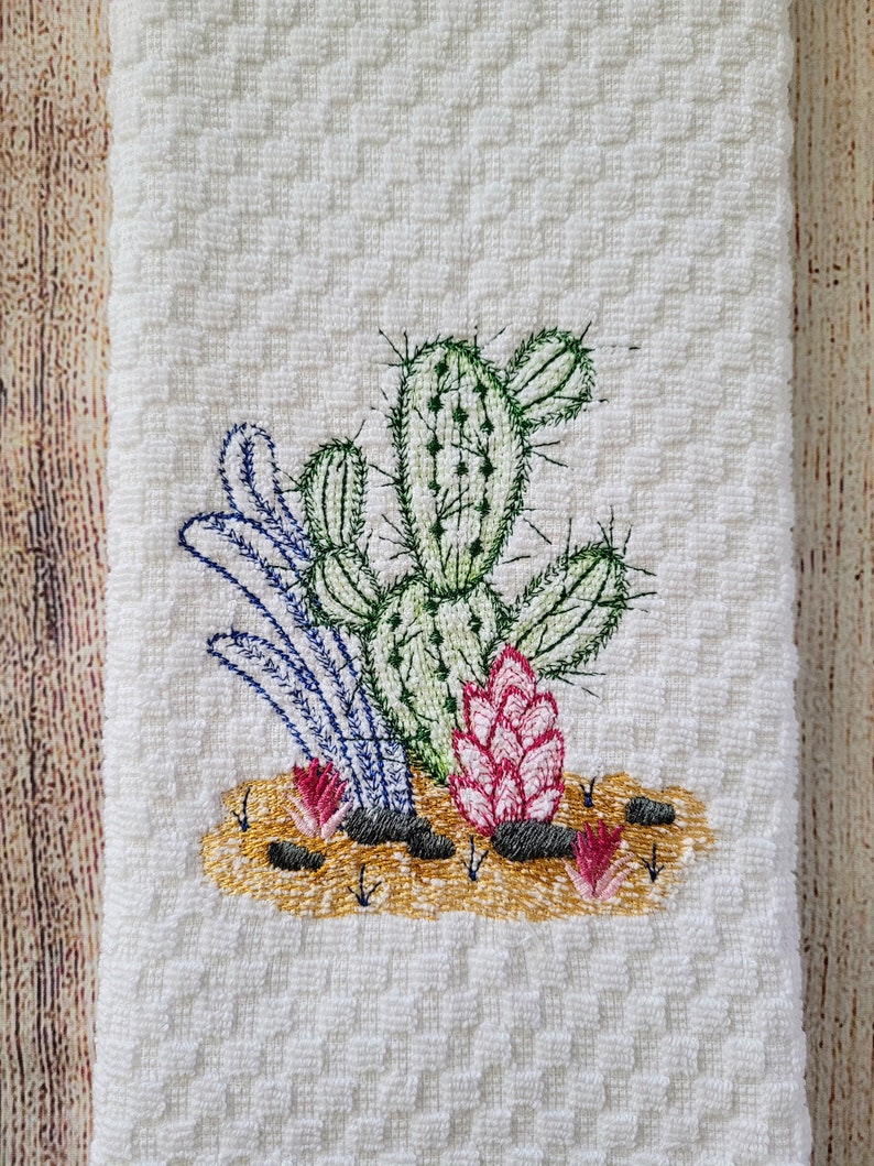 Puede incluir: Toalla de cocina bordada con fondo blanco y un dise&ntilde;o de cactus verde con flores rosas y hojas azules.