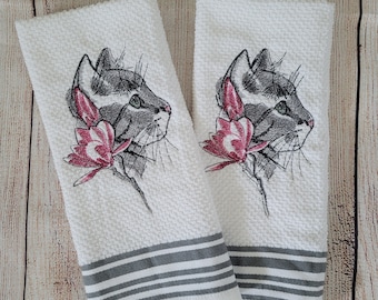 Puede incluir: Dos toallas de cocina blancas con rayas grises. Cada toalla presenta un diseño bordado de la cara de un gato gris con una flor rosa.