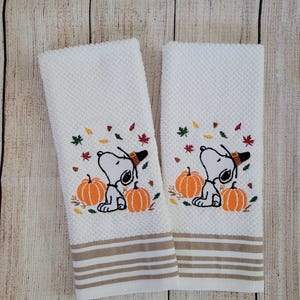 Puede incluir: Dos toallas de cocina blancas con rayas marrones. Cada toalla presenta una imagen bordada de Snoopy con un sombrero de peregrino y sosteniendo una calabaza. El fondo incluye hojas de otoño en tonos naranja, amarillo y rojo.