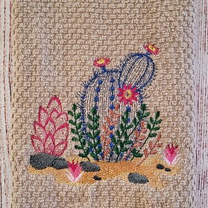 Puede incluir: Toalla de cocina bordada con un dise&ntilde;o de cactus. El cactus es verde con detalles azules y flores rosas. La toalla es beige con textura de tejido de waffle.