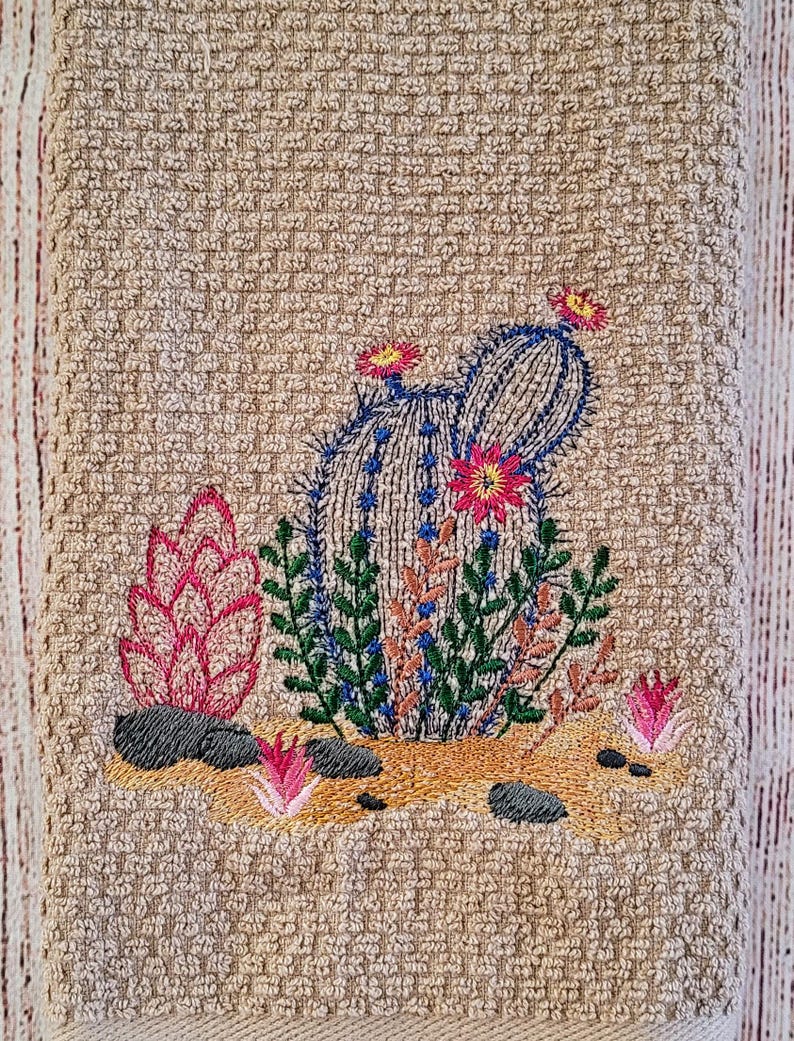 Puede incluir: Toalla de cocina bordada con un dise&ntilde;o de cactus. El cactus es verde con detalles azules y flores rosas. La toalla es beige con textura de tejido de waffle.