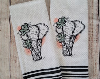 Puede incluir: Dos toallas de cocina blancas con rayas negras. Cada toalla tiene un diseño bordado de un elefante con flores.