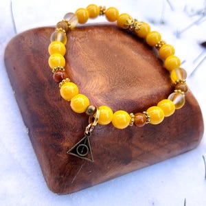 May include: A beaded bracelet featuring large, bright yellow beads interspersed with smaller brown and gold beads. A bronze-colored charm in the shape of a triangle with a circle inside hangs from the bracelet. The bracelet is displayed on a wooden surface.