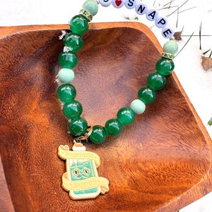 May include: A beaded bracelet with green beads and a gold-colored charm. The charm features a potion bottle design with the words "Potions" and "Potion No." The bracelet also spells out "S ❤️ Snape" with letter beads.