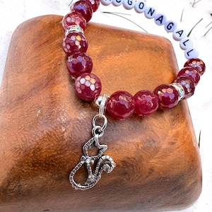 May include: A burgundy beaded bracelet with the name "MCGONAGALL" spelled out in white letter beads. The bracelet features a silver charm of a cat. The beads have a textured, faceted appearance.